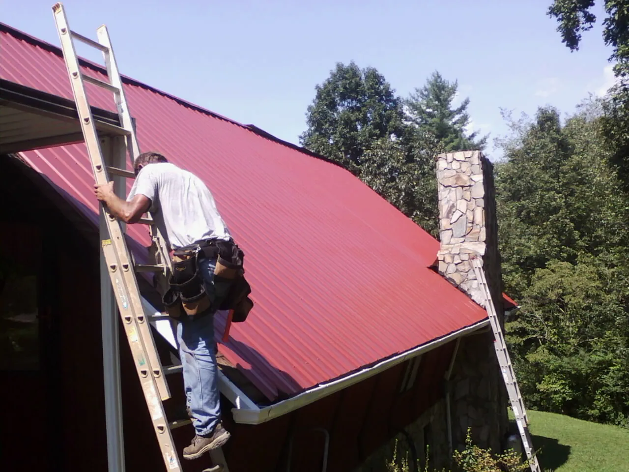 Roofer on a ladder beside a red metal roof and stone chimney — Soffit & Fascia Repair by Roofing Brothers in Kerman